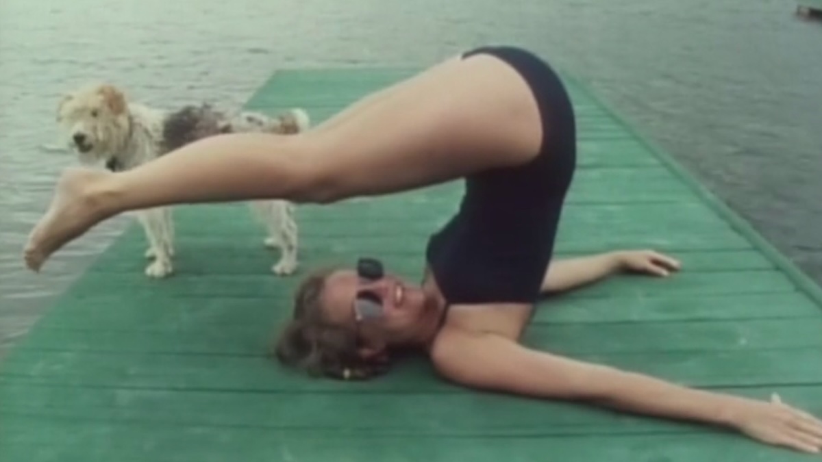 Film still of a woman on a boat dock doing an acrobatic pose. She is wearing a black bathing suit and sunglasses. A dog is standing behind her.