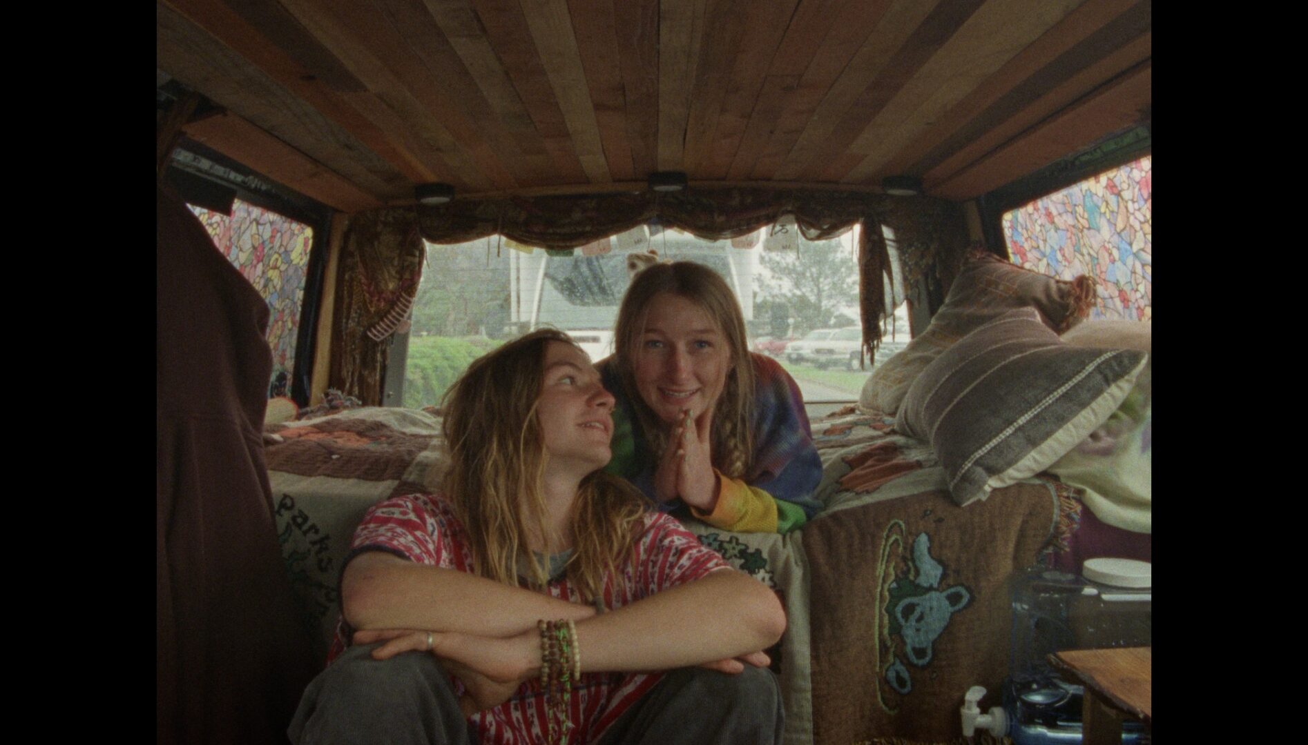 Film still of two young adults with long blonde hair and tie dyed shirts in the back of a van.