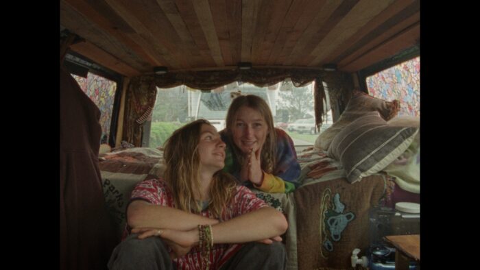 Film still of two young adults with long blonde hair and tie dyed shirts in the back of a van.