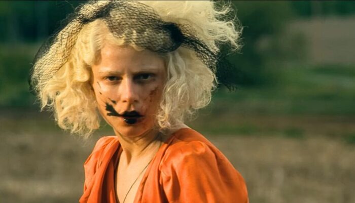 Film still of a woman with curly, light blonde hair, black smeared around her mouth, wearing an orange dress.