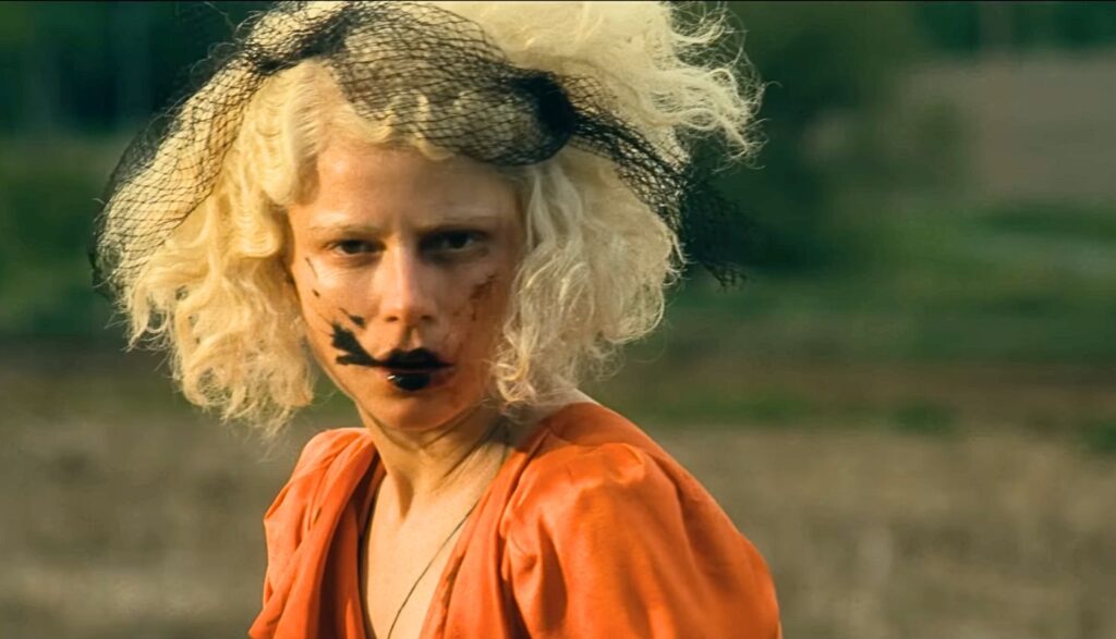Film still of a woman with curly, light blonde hair, black smeared around her mouth, wearing an orange dress.
