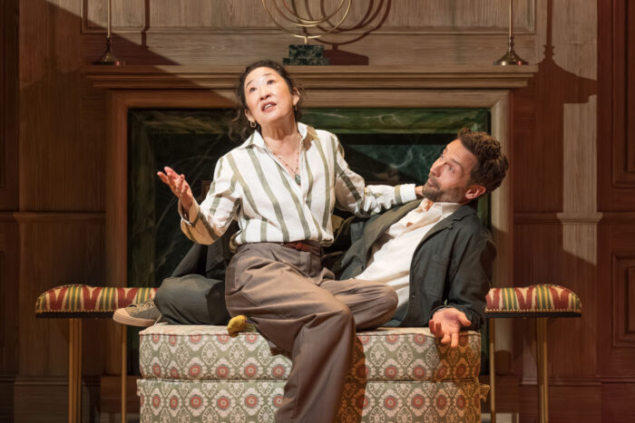 Photo of two actors on stage, a woman and a man sitting on a couch.
