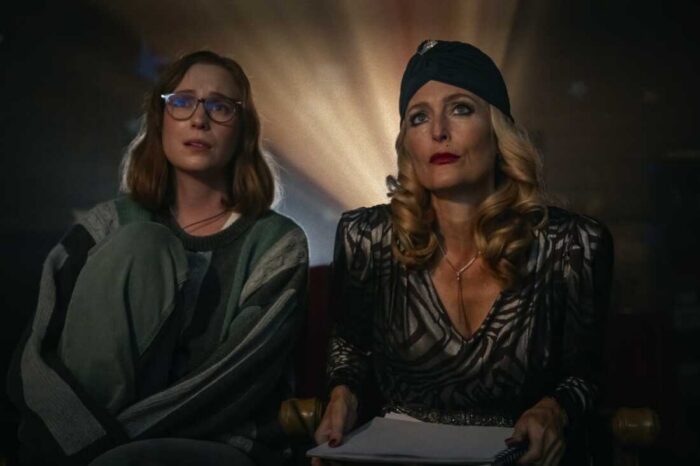 Film still of two women sitting looking forward with a movie camera light behind them.