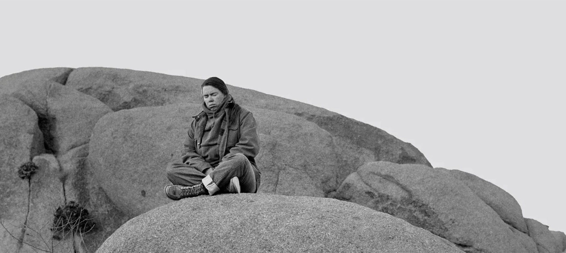 Black and white photo of a woman sitting cross-legged on a boulder with her eyes closed and head bowed.