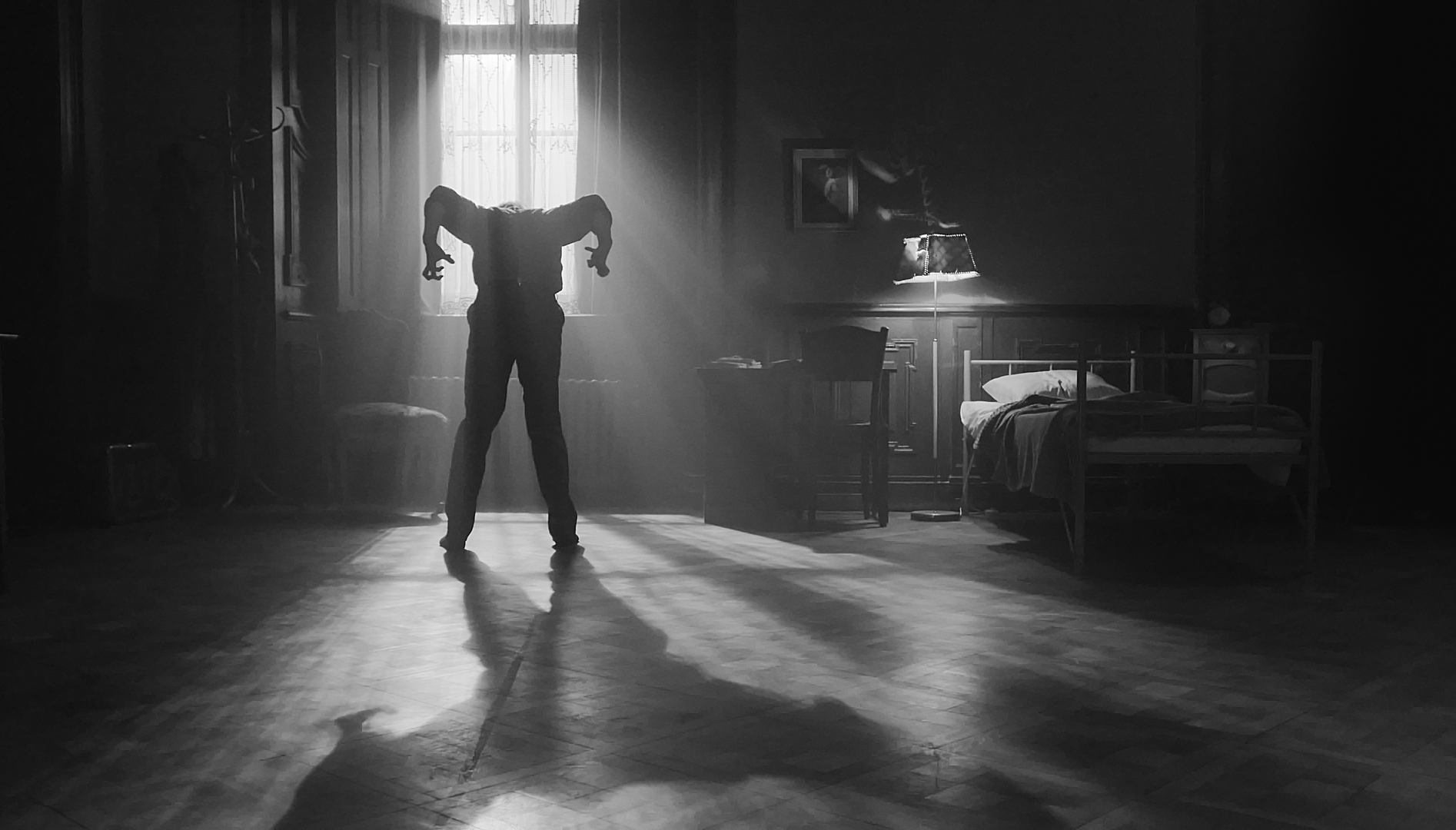 Black and white film still of a dim room with light only coming from one window. In front of the window a person is standing with their head down and their elbows up in the air.