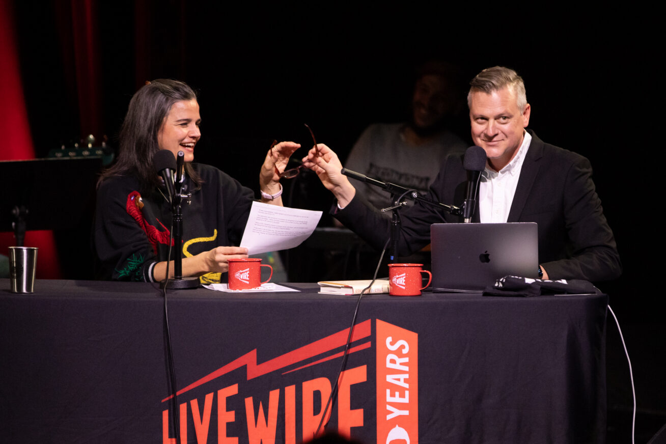 A man and woman on stage at a table with microphones and a banner that says Live Wire.