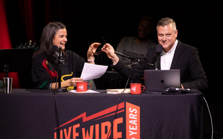 A man and woman on stage at a table with microphones and a banner that says Live Wire.