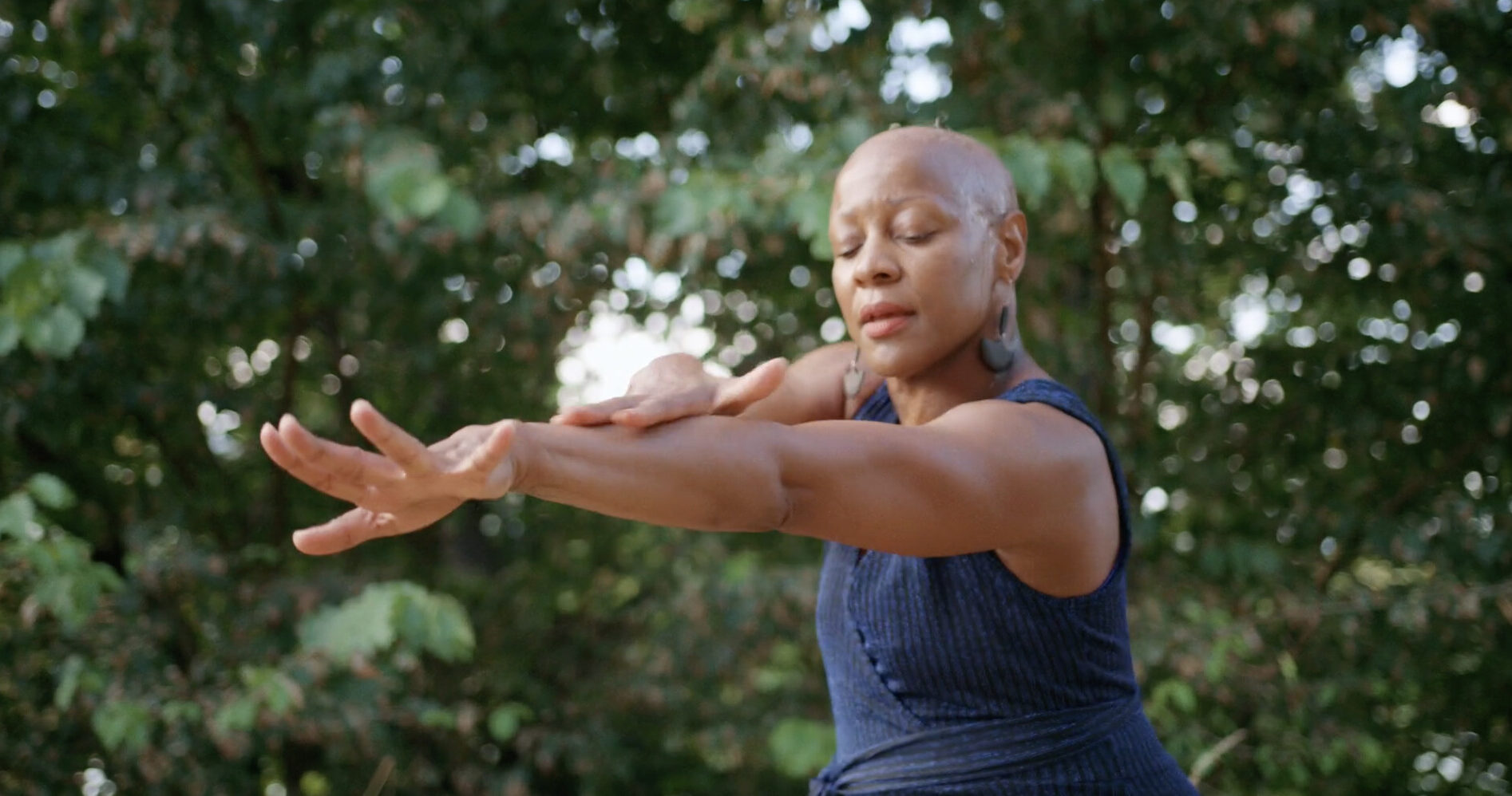 Photo of a Black dancer with their eyes closed, holding one arm forward and the other bent over the outstretched arm.