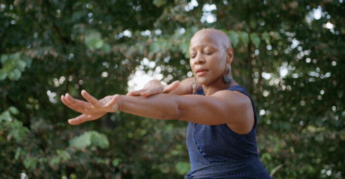 Photo of a Black dancer with their eyes closed, holding one arm forward and the other bent over the outstretched arm.