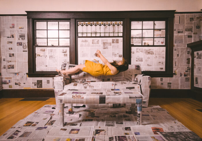 Film still of a person levitating above a couch that is covered with newspapers, in a room covered in newspapers.