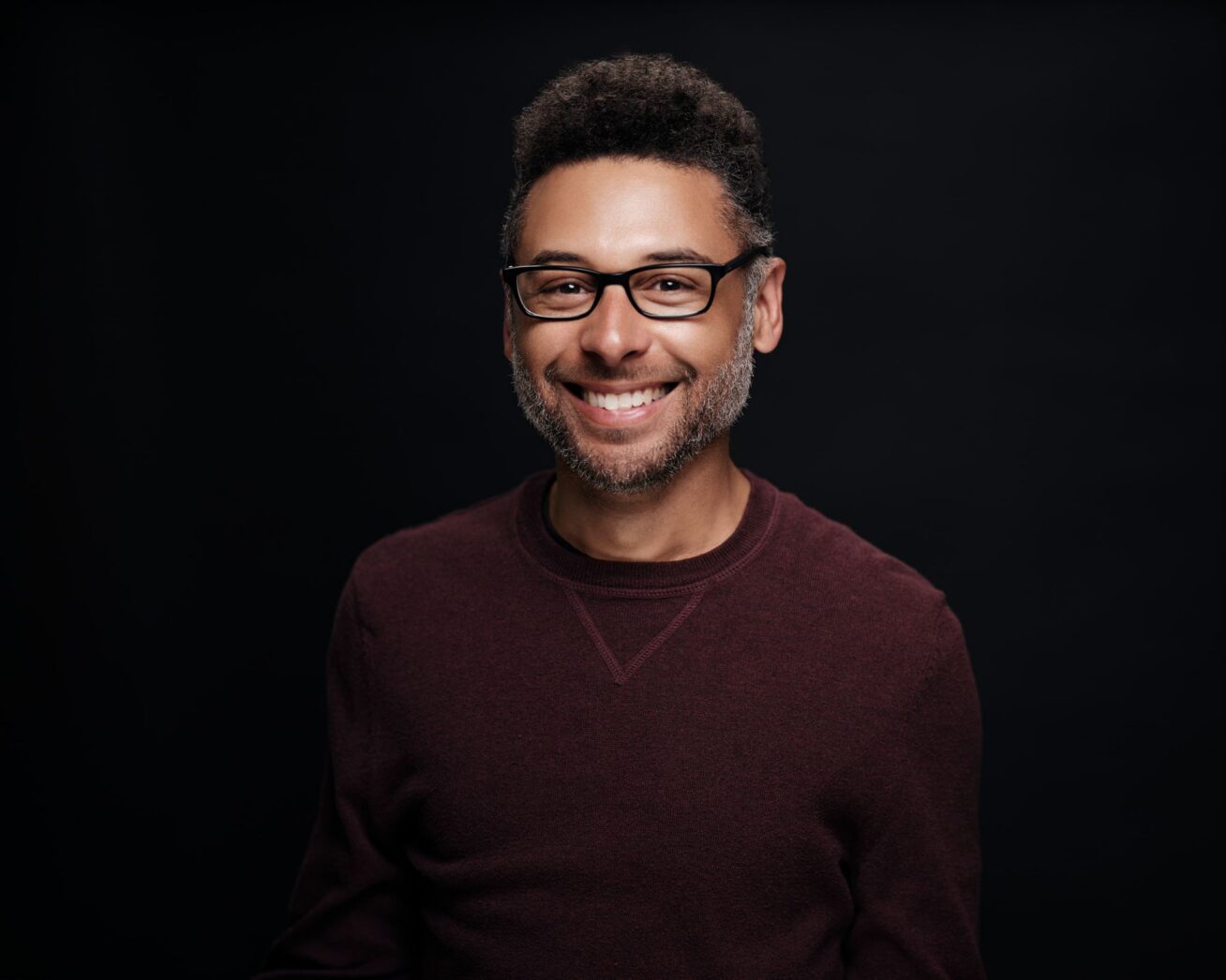 Photo of a smiling Black man wearing  black eyeglasses and a maroon sweater.