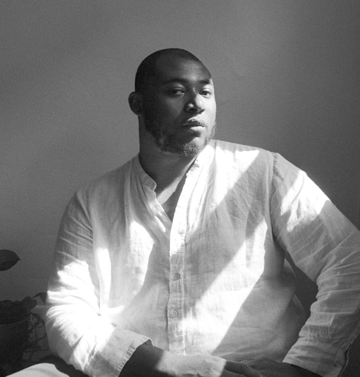 Black and white photo of a Black man in a white button down shirt, sitting in front of a window and partially lit by the sunlight.