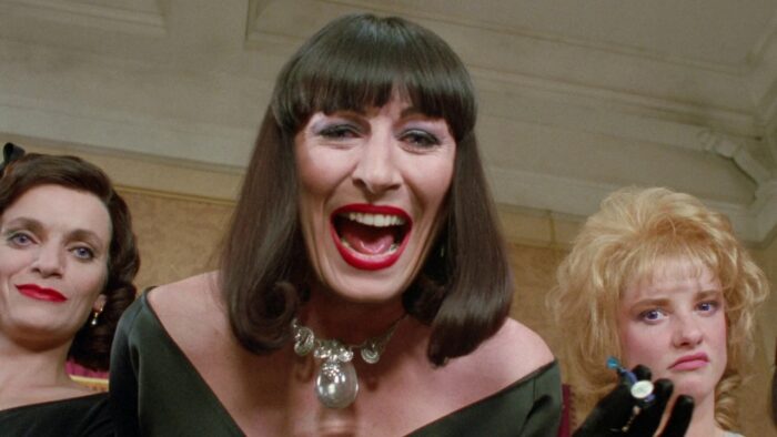 Film still of a Anjelica Huston yelling towards the camera. Two women stand to her sides.
