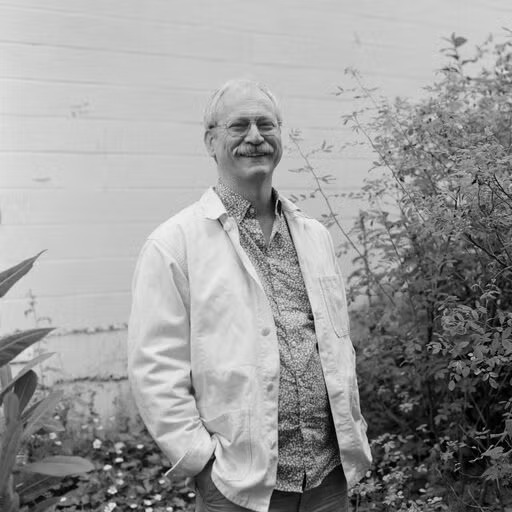 Black and white photo of a white man in a white jacket with his hands in his pockets.