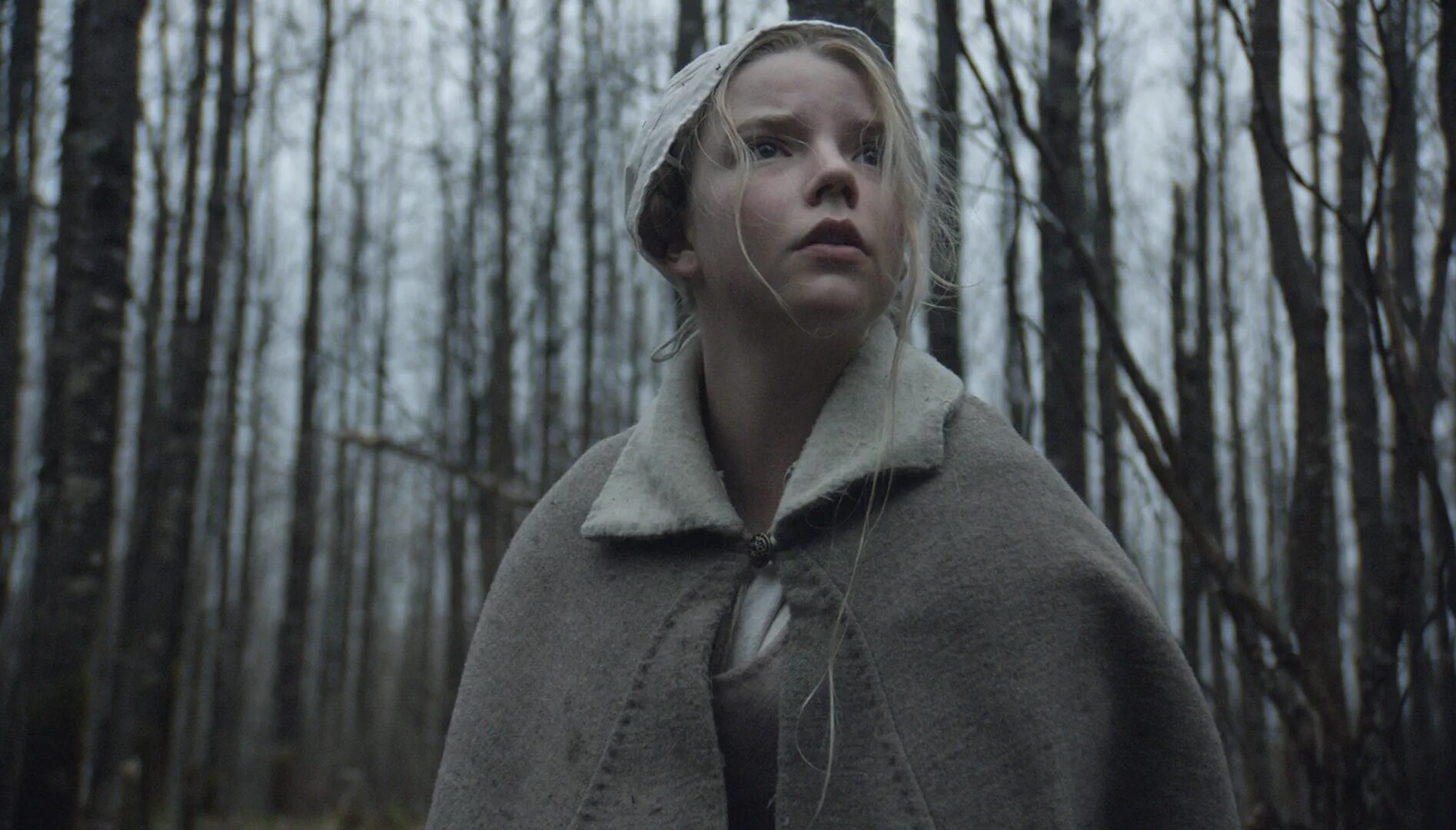 Film still of a blond girl in a cap standing with bare trees behind her with a scared look on her face.