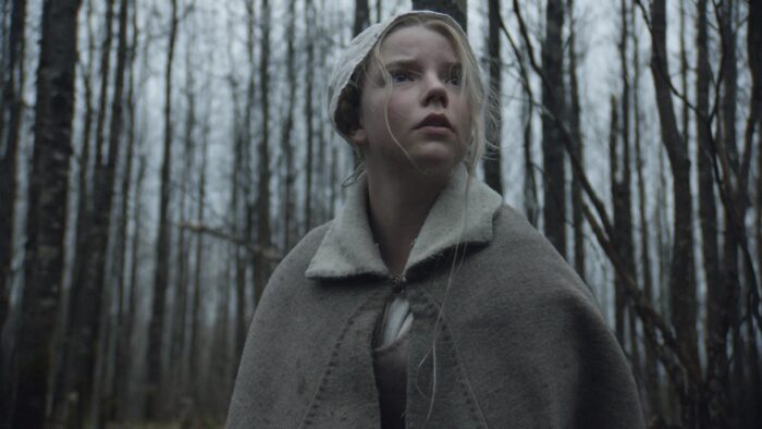 Film still of a blond girl in a cap standing with bare trees behind her with a scared look on her face.