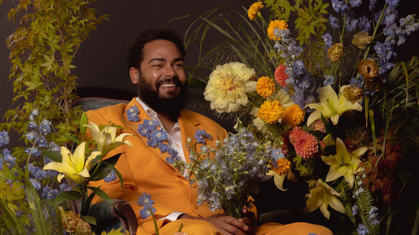 Image of a smiling Black man in an orange suit, surrounded by flowers.