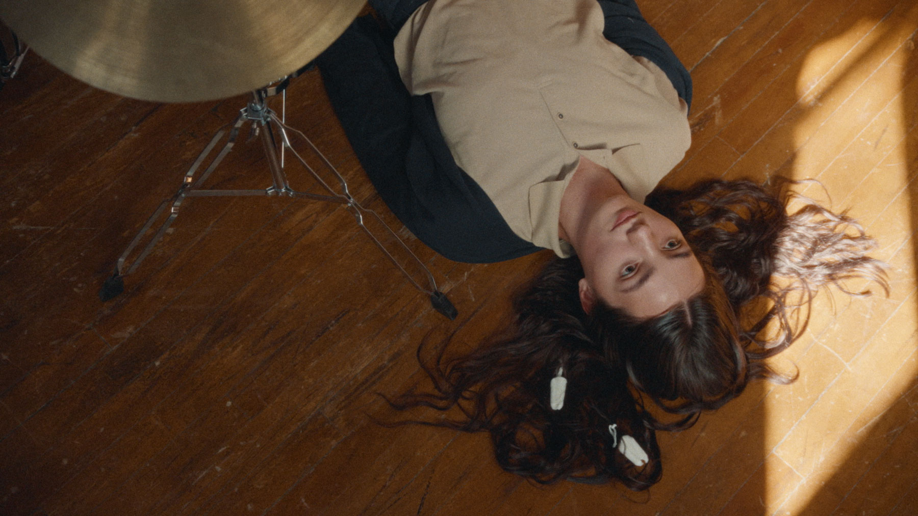 A young woman laying on a wood floor below a drum set, with her hair spread out around her head.
