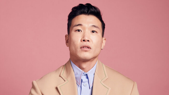 Photo of Joel Kim Booster, wearing a tan jacker, a blue collared shirt, and sitting against a pink background.