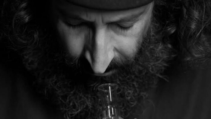 Black and white film still of a man with long hair and a beard smelling a vial of perfume.