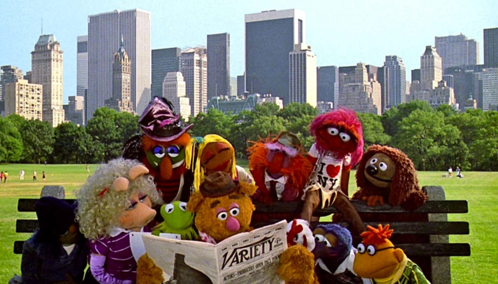 Film still of the Muppets on a park bench with city buildings behind them.