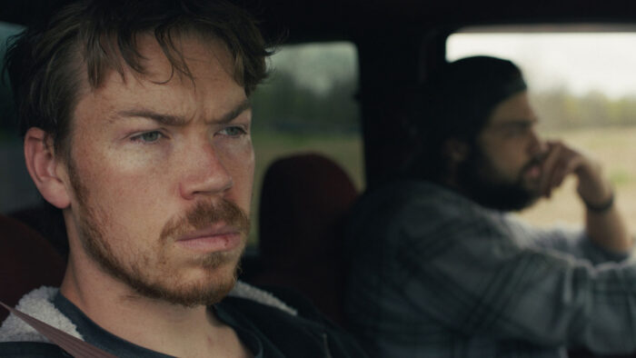 Film still of a pensive looking man in the passenger seat of a vehicle.